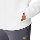 A person wears the Everest Training Full-Zip Hoodie by Canterbury in white, with one hand in the pocket, paired with gray pants featuring a yellow Canterbury logo.