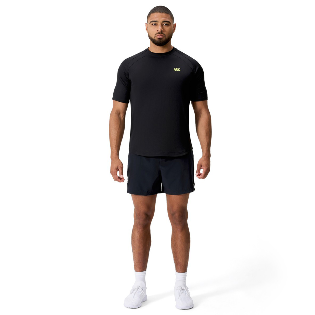 A man faces forward against a white background, wearing the Canterbury Everest Training Tee in black, paired with black shorts and white athletic shoes.