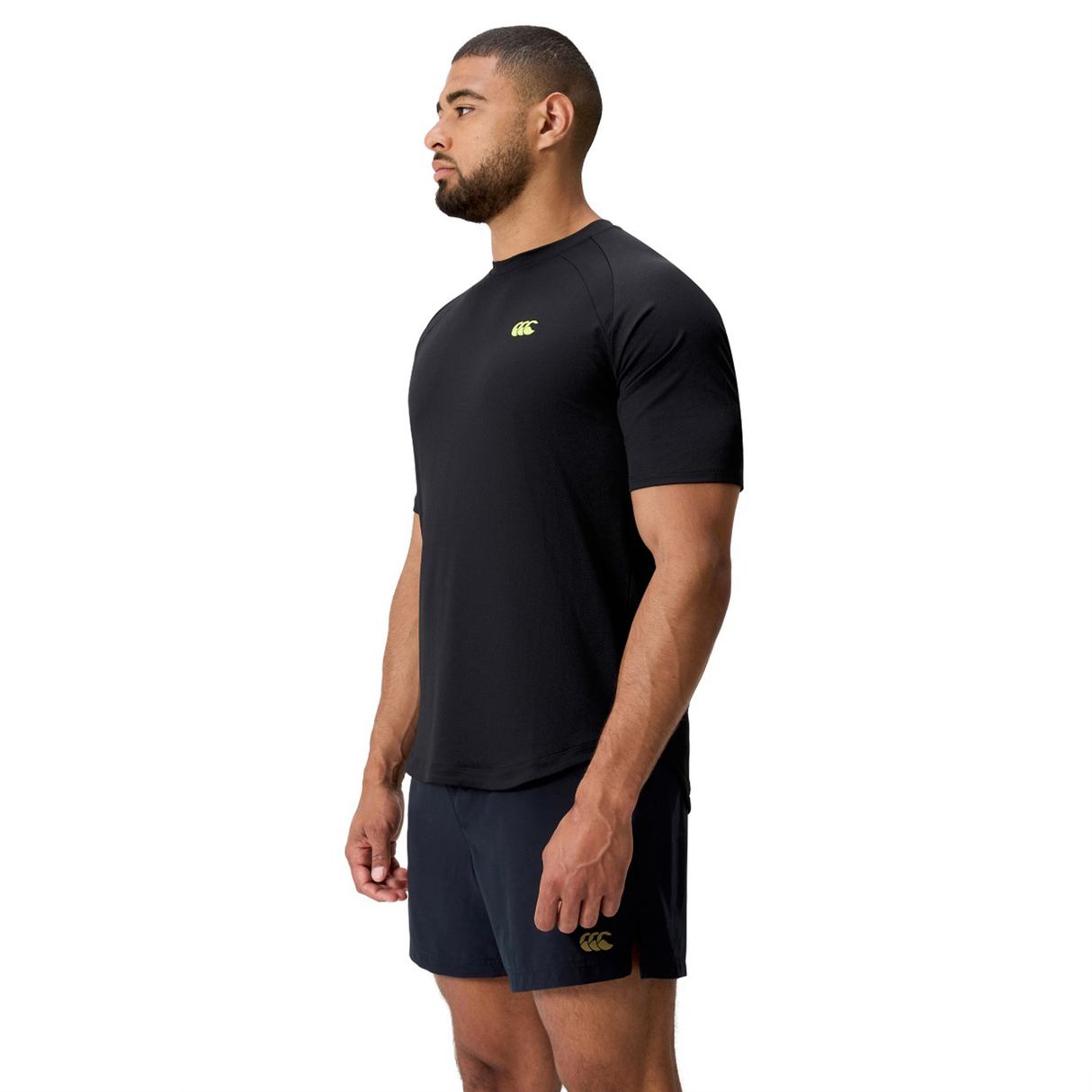A man stands in side profile, wearing the black Everest Training Tee by Canterbury and dark shorts against a white background—ideal for rugby players who want both style and comfort.
