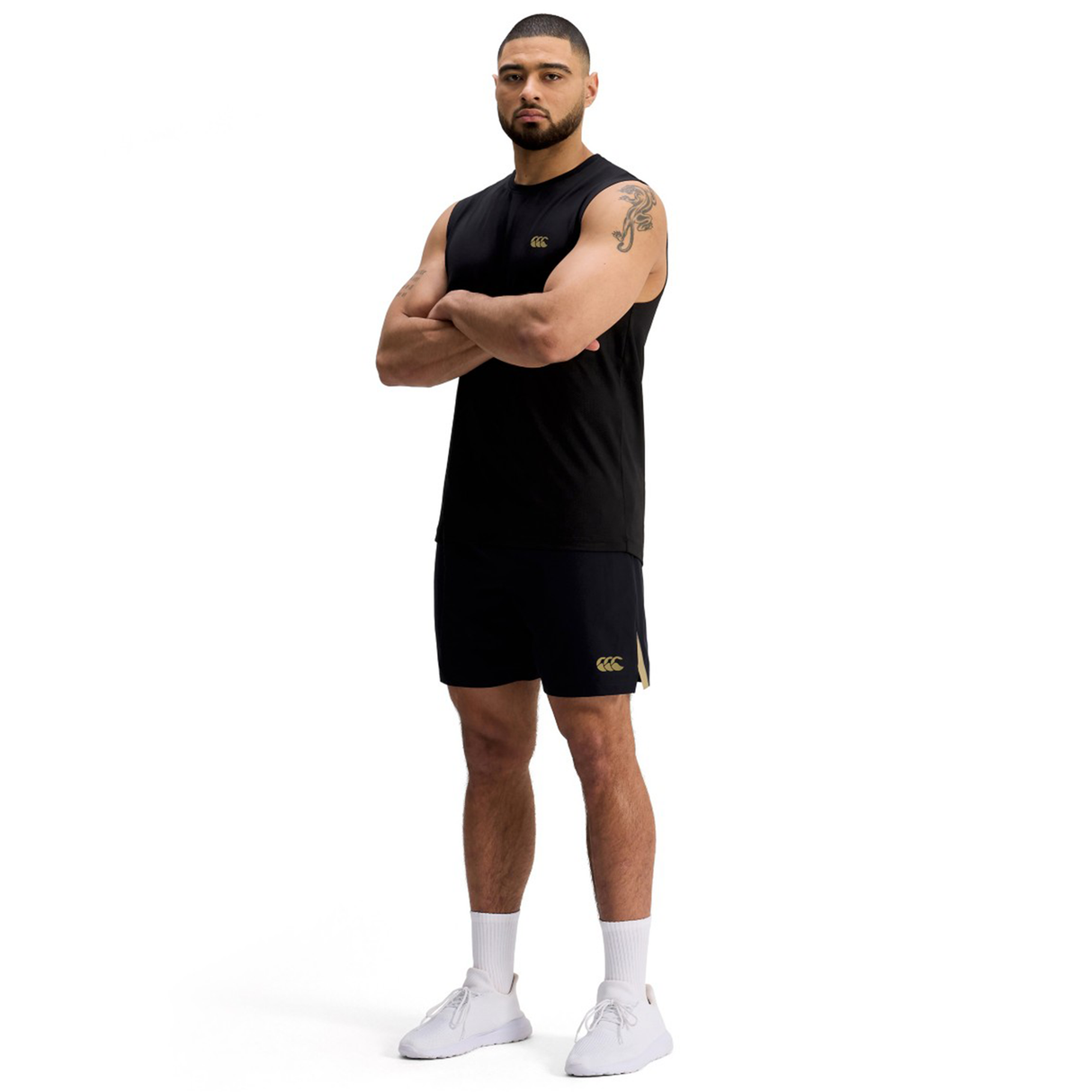 A man stands with arms crossed, wearing black Canterbury Tempo+ 7" Training Shorts, a black sleeveless shirt, white socks, and white athletic shoes against a plain white background.