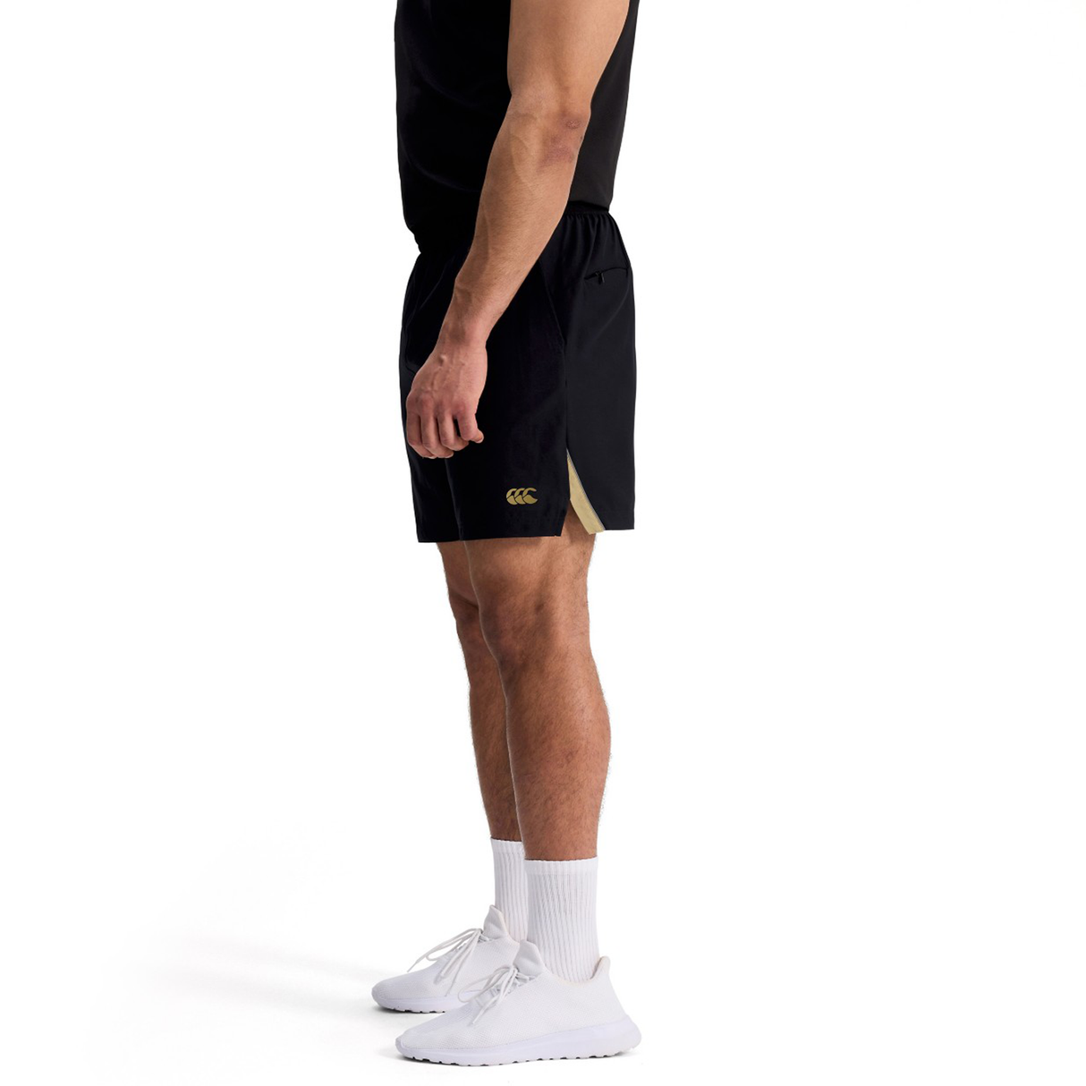 A man in a black sleeveless shirt, Canterbury Tempo+ 7" Training Shorts, white socks, and white sneakers stands in profile against a white background.