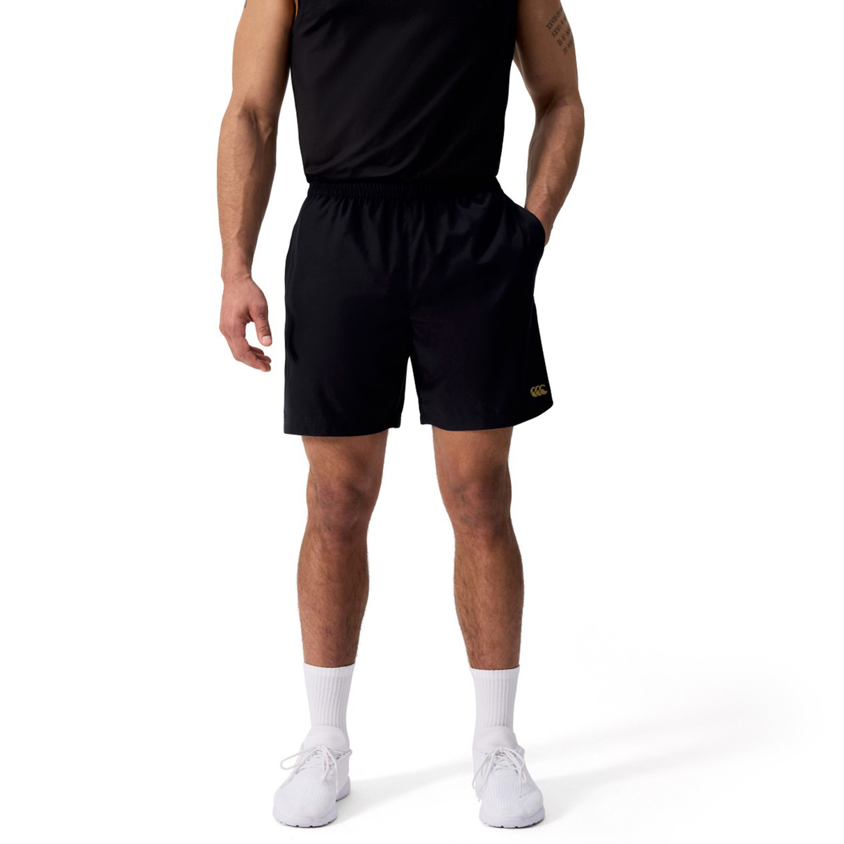 A person wears black Canterbury Tempo+ 7" Training Shorts, an athletic shirt, white socks, and white sneakers, standing with hands in pockets against a plain background.
