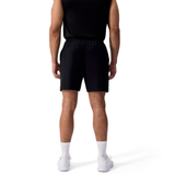 A man faces away, wearing a black sleeveless shirt and Canterbury Tempo+ 7" Training Shorts, paired with white socks and sneakers, against a plain white background.