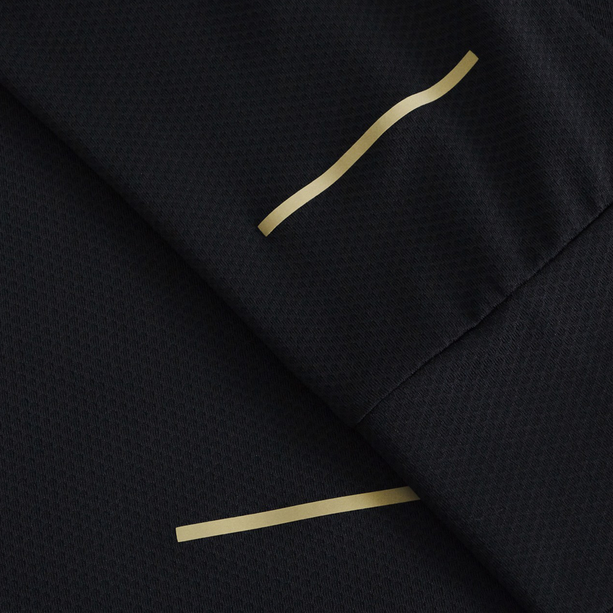 Close-up of black textured fabric from the Canterbury Tempo+ 1/4 Zip Training Top, showing two thin gold-colored lines—one straight, one wavy—across overlapping folds.