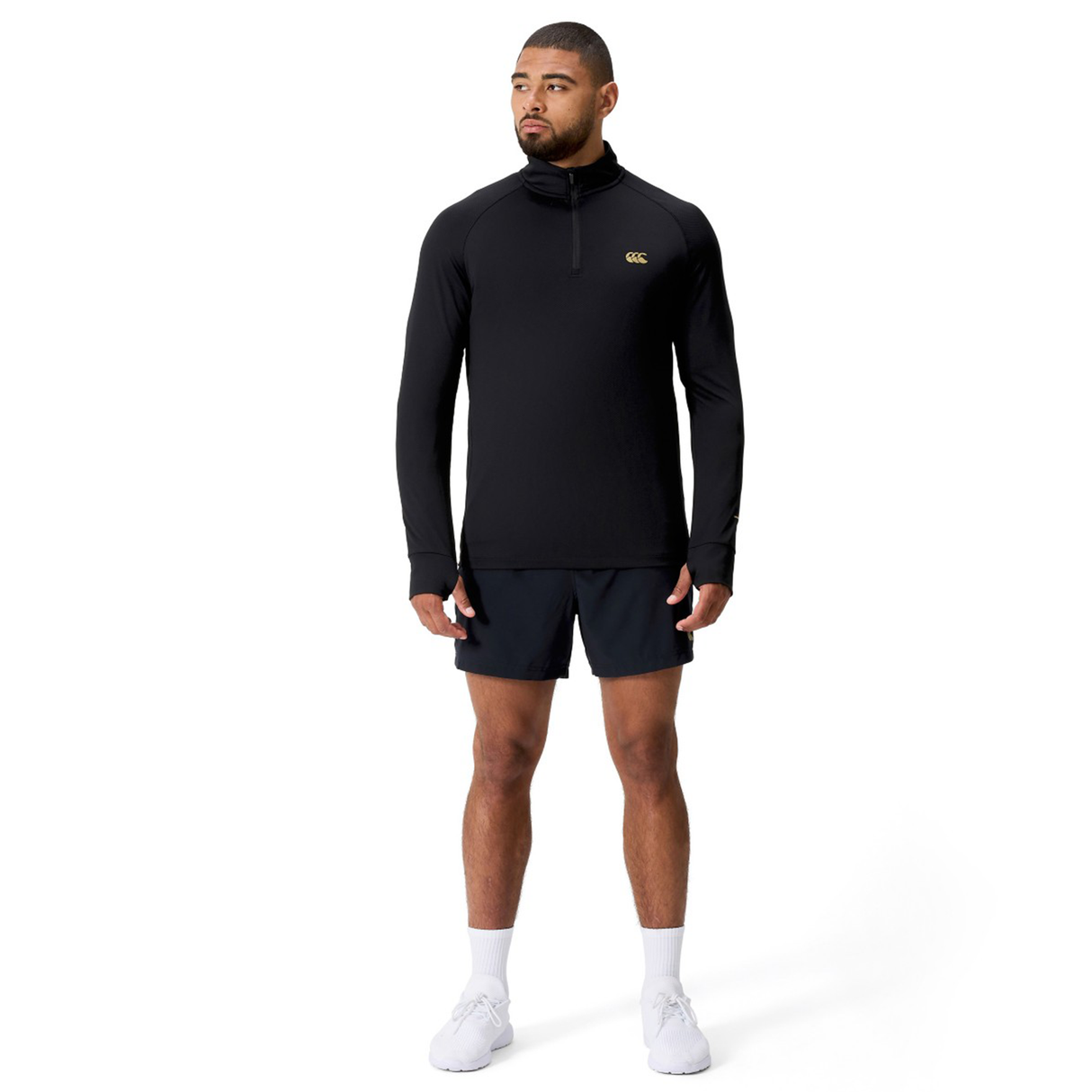 A man faces slightly to the side, wearing a black Canterbury Tempo+ 1/4 Zip Training Top, black shorts, white socks, and white sneakers against a plain white background.
