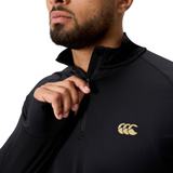 A person zips up the collar of a black Canterbury Tempo+ 1/4 Zip Training Top featuring a gold sports logo on the chest.