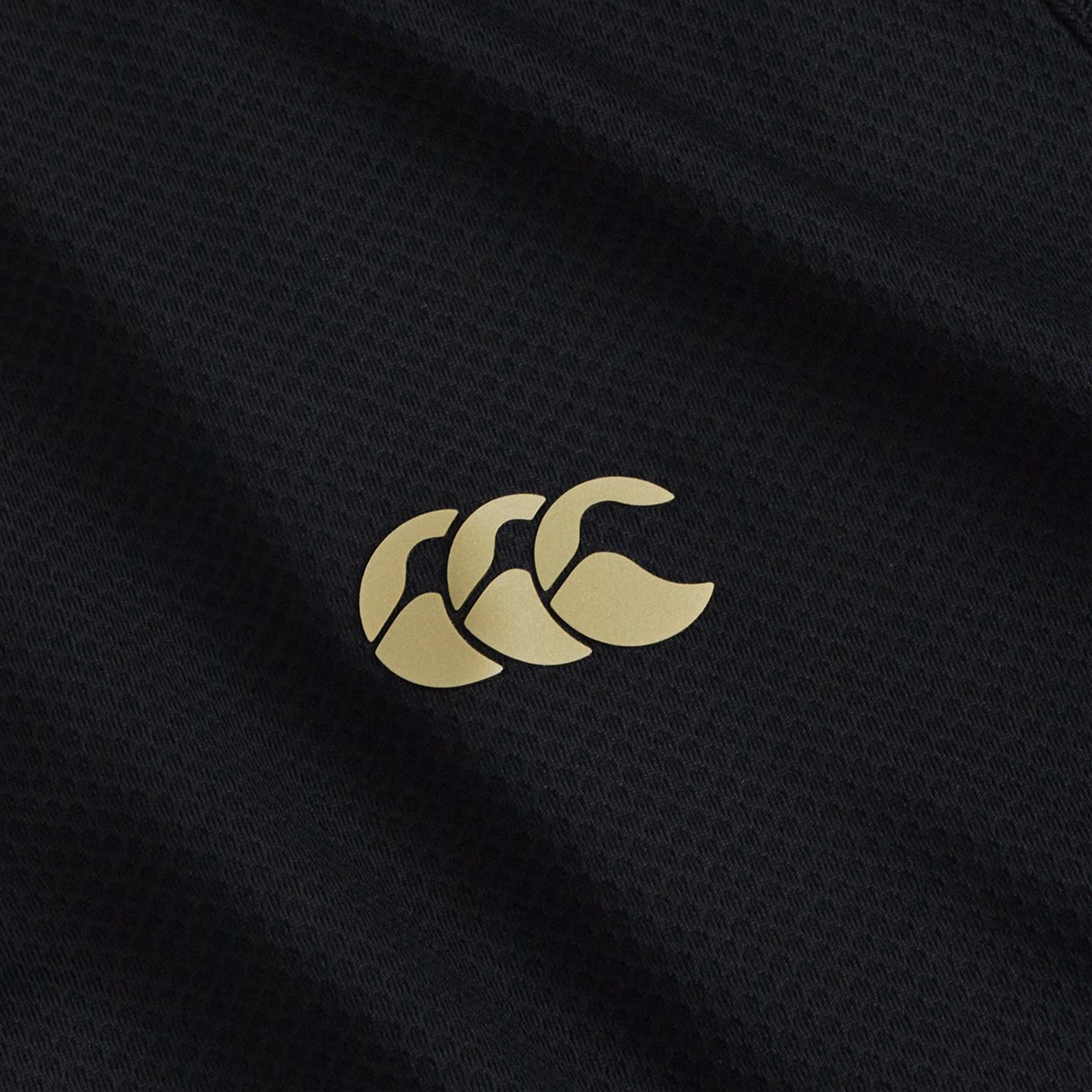 Close-up of the black Tempo+ 1/4 Zip Training Top by Canterbury, featuring a gold logo with three overlapping stylized “C” shapes—a lightweight performance fabric ideal for rugby training.
