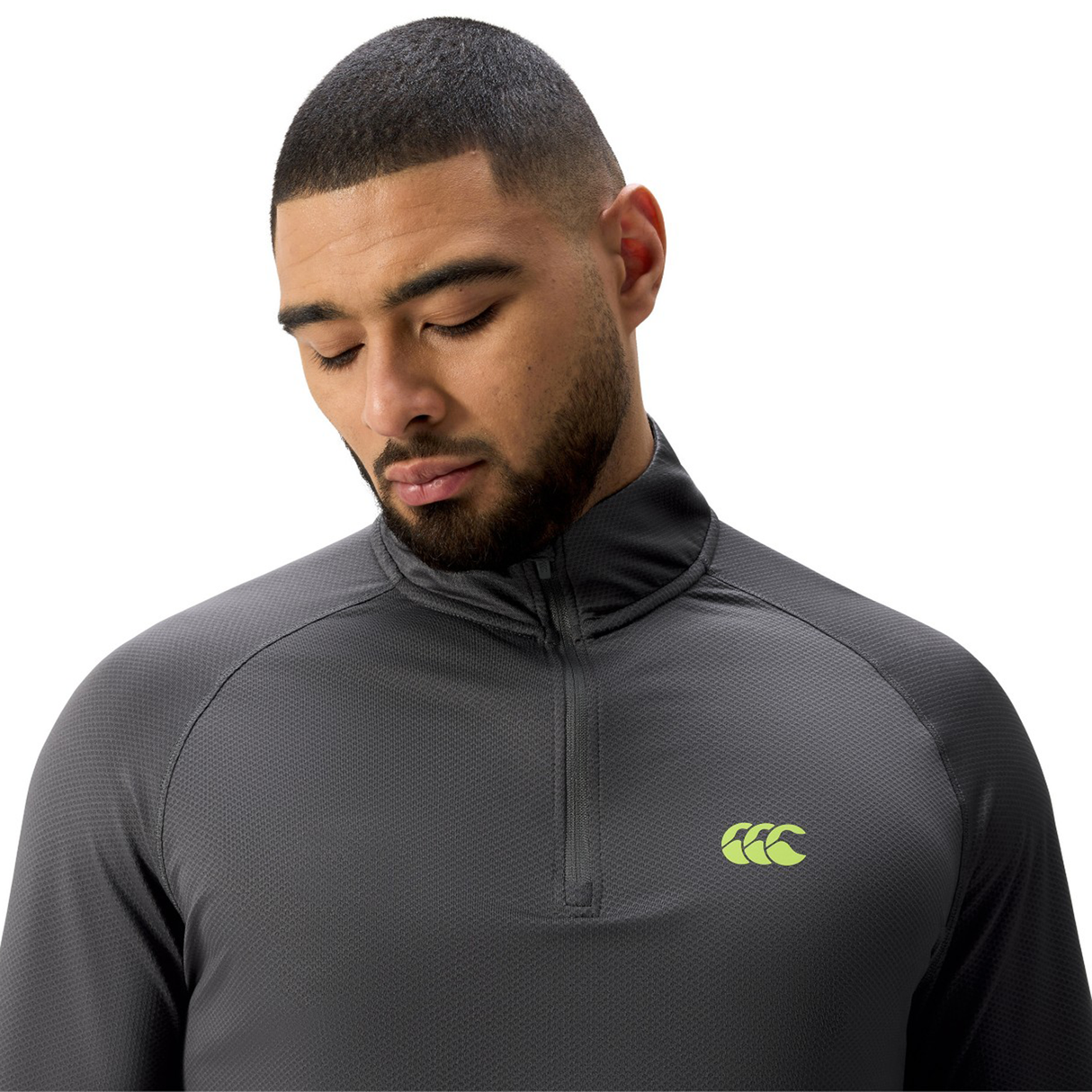 A man with short hair and a beard wears a dark gray Canterbury Tempo+ 1/4 Zip Training Top featuring a yellow logo, looking down against a white background.