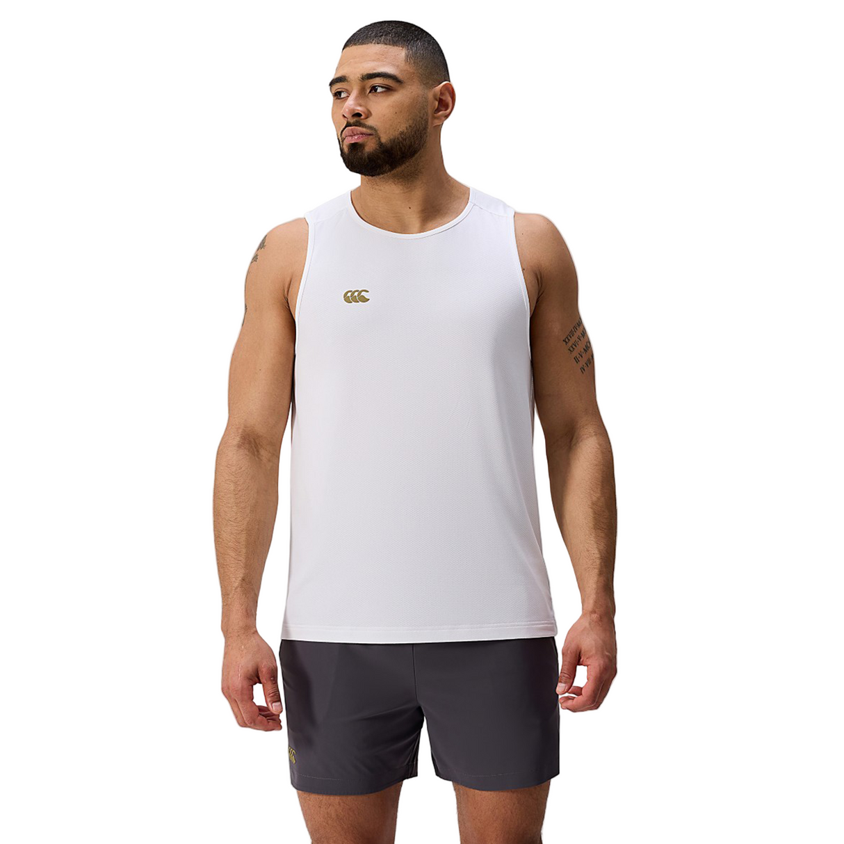 A man stands facing forward against a plain light background, wearing the Canterbury Tempo+ Singlet, a white moisture-wicking gym top, paired with dark shorts.