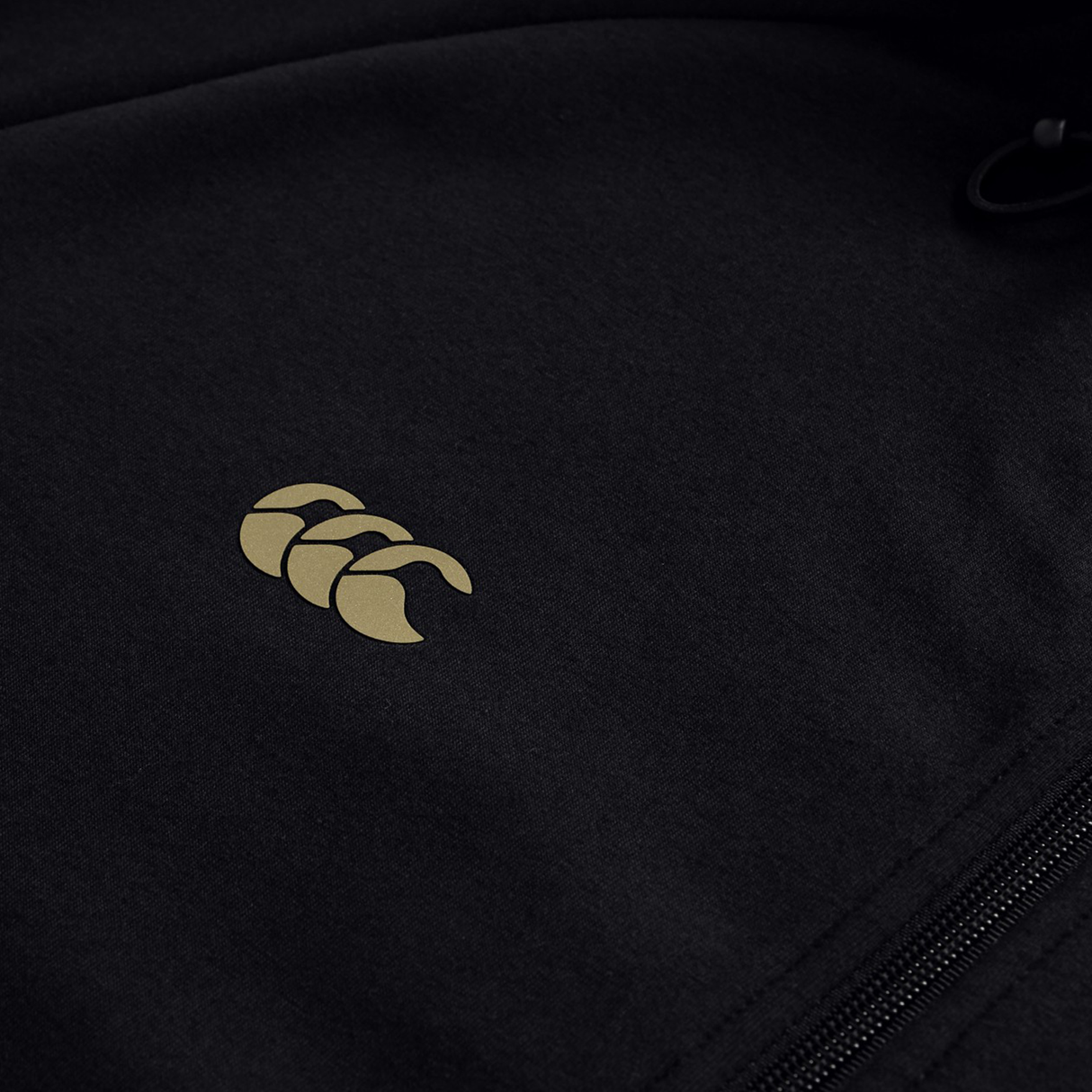 Close-up of the Canterbury Tempo+ Full-Zip Hoodie by Canterbury, a black lightweight stretch hoodie featuring a small gold logo with three overlapping shapes and a visible right-side zipper.