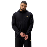 A man stands against a plain white background wearing the Canterbury Tempo+ Full-Zip Hoodie by Canterbury in black, featuring a hood and gold logo, paired with matching black pants.