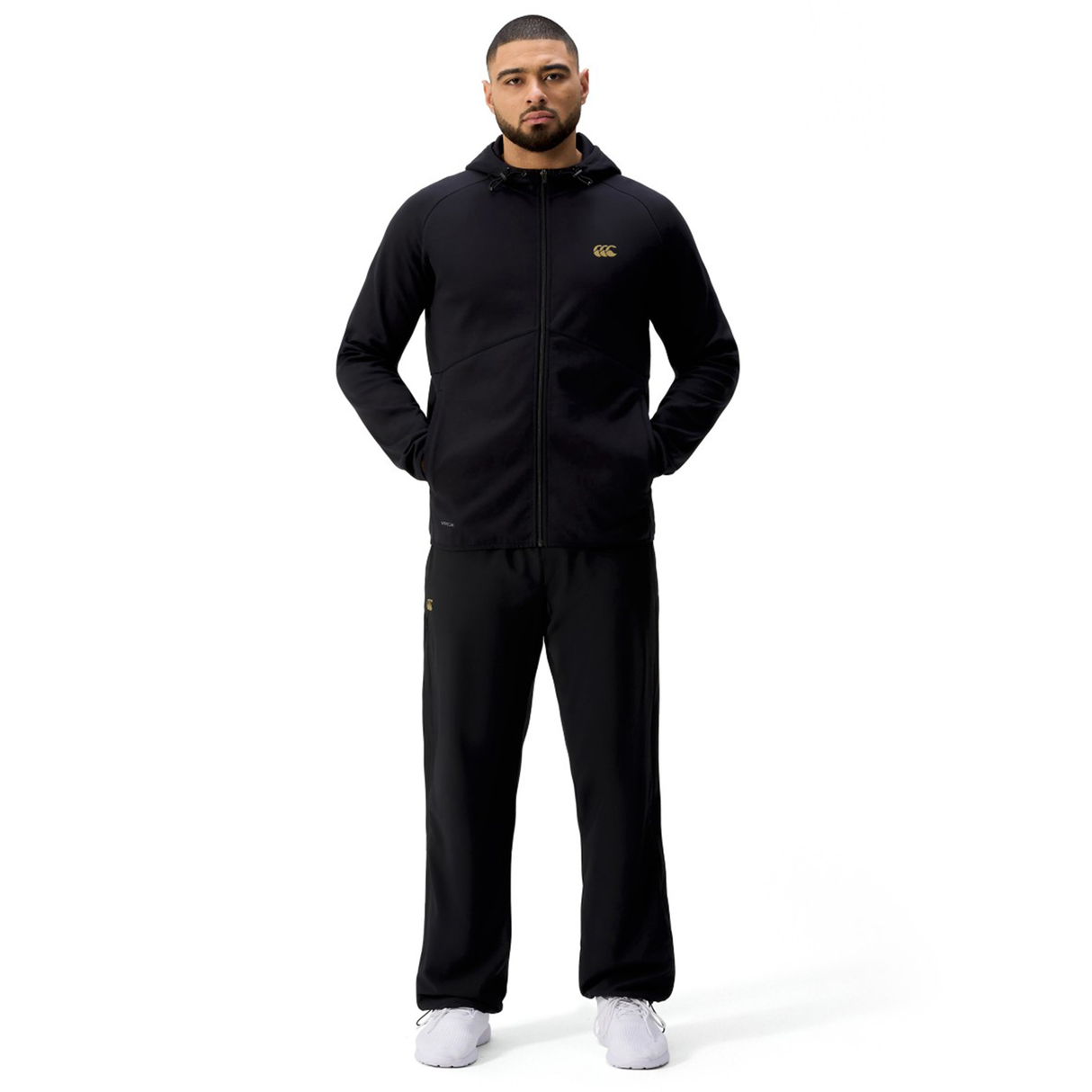 A man stands facing forward against a plain white background, wearing a Canterbury Tempo+ Full-Zip Hoodie in black, paired with black pants and white sneakers.