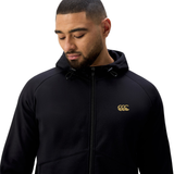A man wearing a black Canterbury Tempo+ Full-Zip Hoodie by Canterbury, featuring a gold logo on the left chest, looks down against a white background.