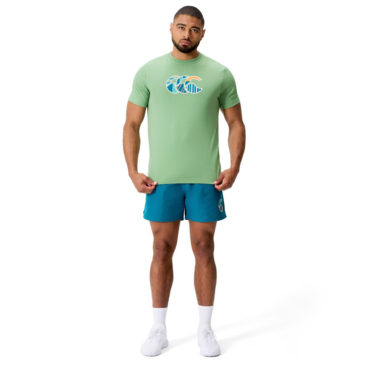 Man against white background wears a light green Canterbury Uglies CCC Logo T-Shirt by Canterbury H225, teal shorts, white socks, and white sneakers.