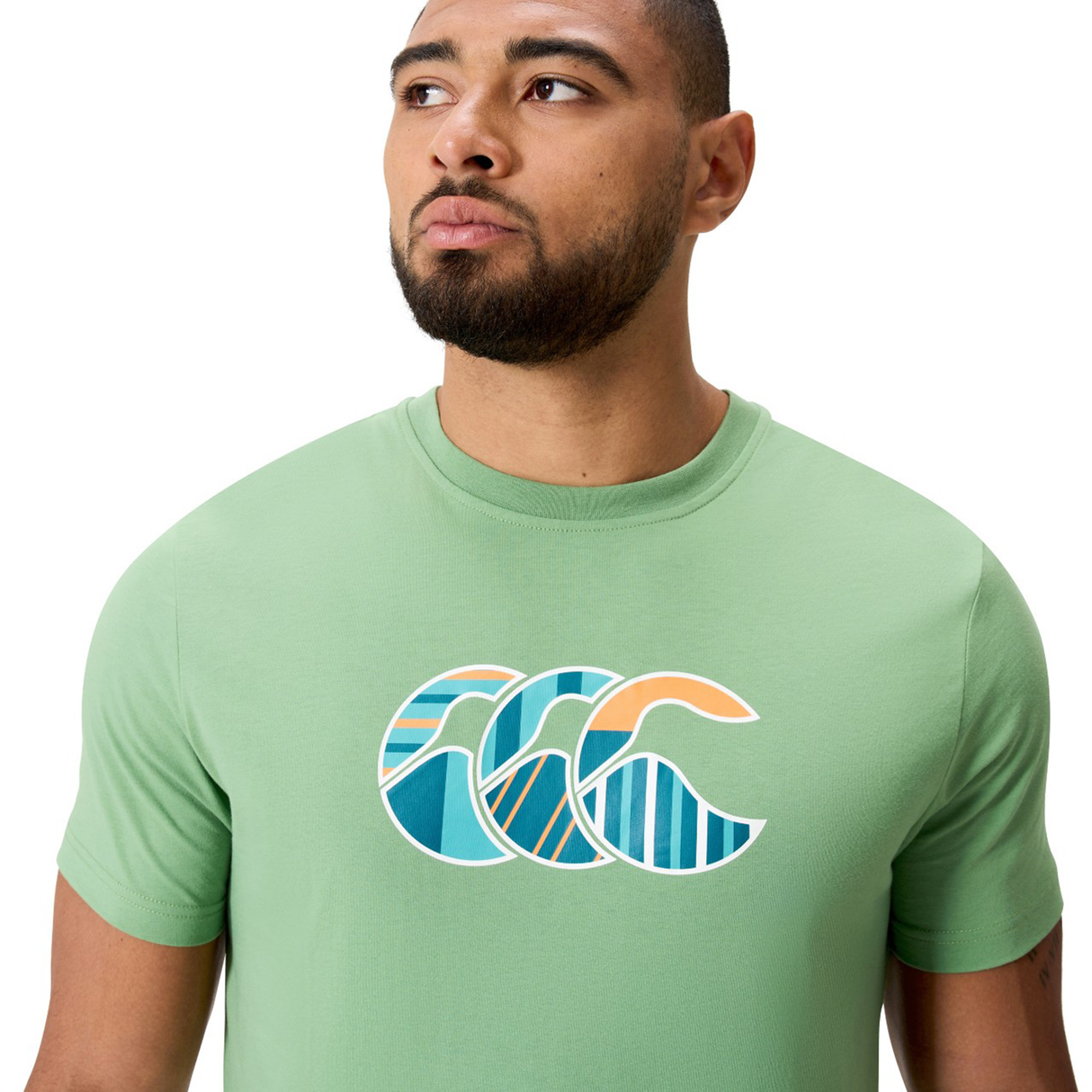A person wearing a light green Canterbury Uglies CCC Logo T-Shirt by Canterbury H225, featuring a graphic of three stylized ocean waves in blue, teal, and orange.