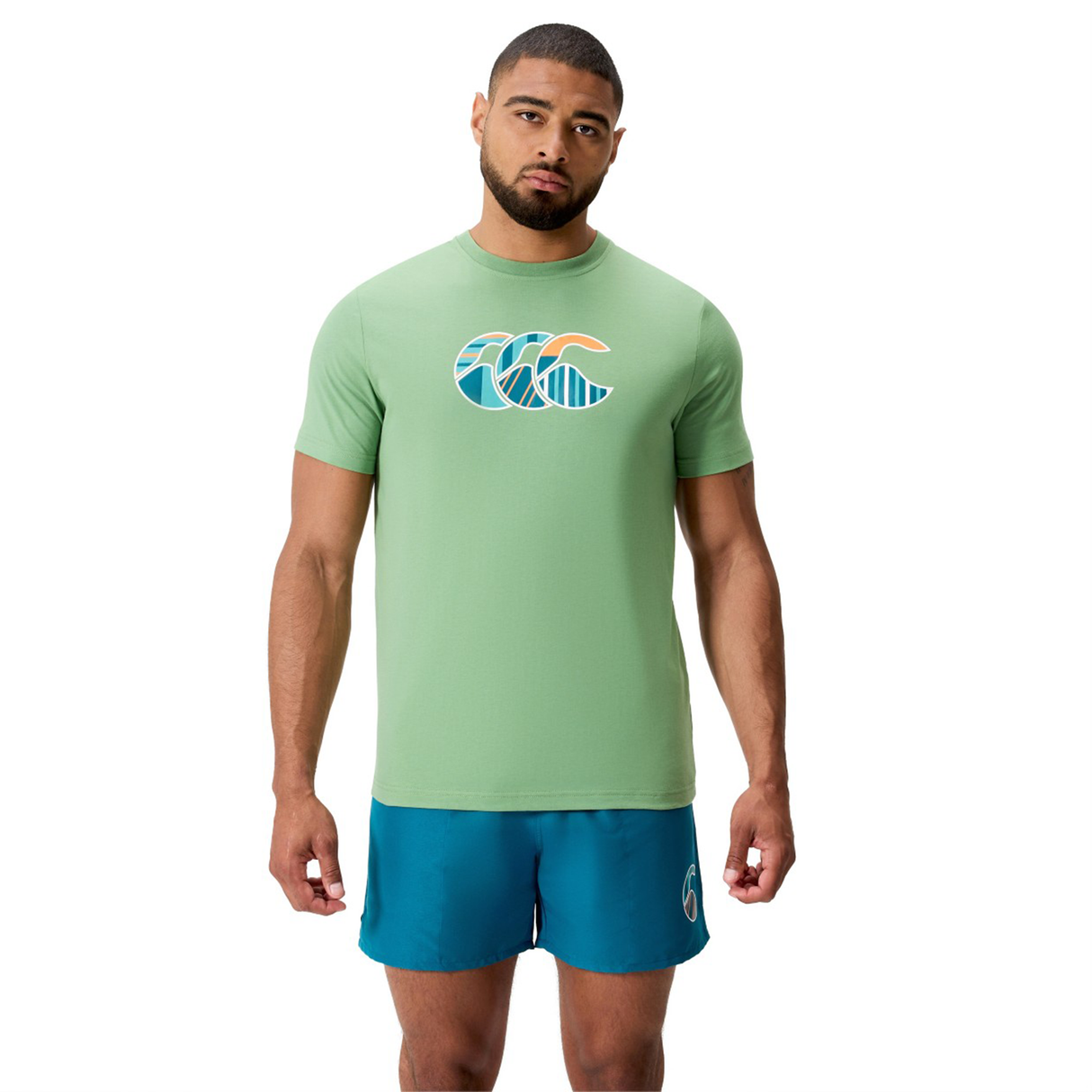 A man wearing a light green Canterbury Uglies CCC Logo T-Shirt by Canterbury H225 and teal athletic shorts stands facing forward against a plain white background.