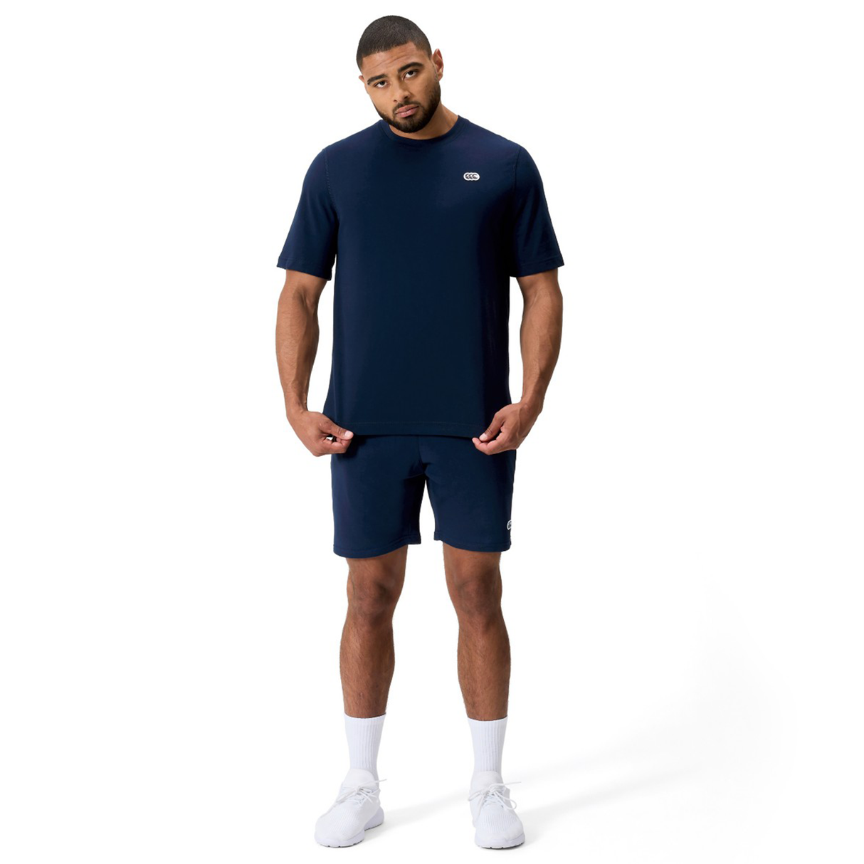 A man stands facing forward wearing a navy blue Canterbury Sport Dept. Logo T-Shirt, matching shorts, white socks, and white sneakers against a plain white background.