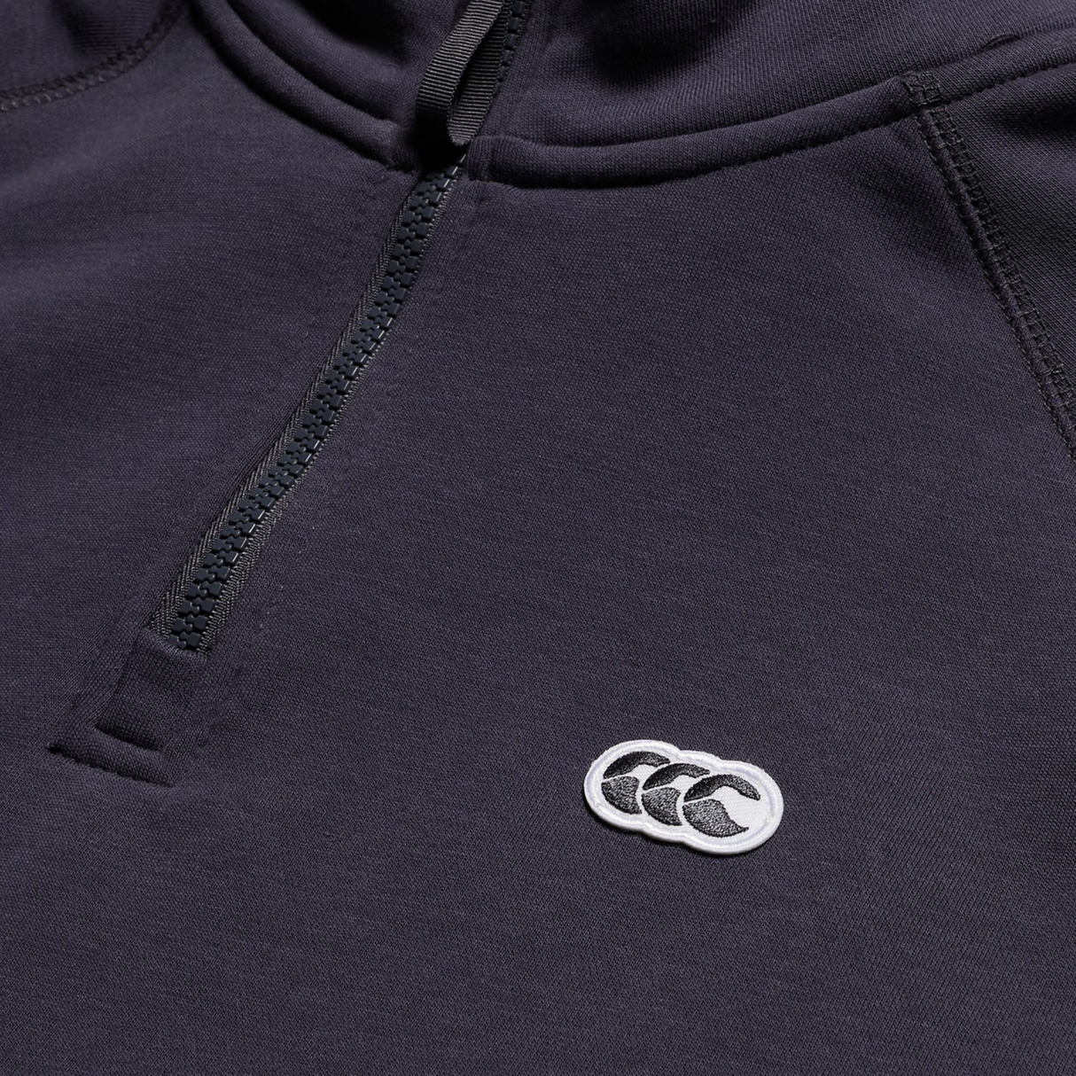 Close-up of the Canterbury Sport Dept. 1/4 Zip Sweater in a dark shade, showing a small white chest logo with three overlapping shapes.