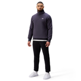 A man stands facing forward against a white background, wearing a Canterbury Sport Dept. 1/4 Zip Sweater, black pants, a white shirt underneath, and white sneakers.