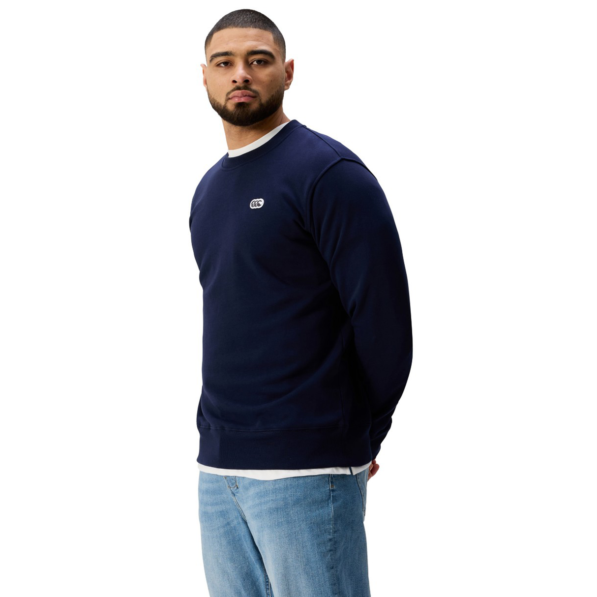 A man stands slightly turned against a white background, wearing Canterbury's Sport Dept. Crew Sweatshirt in navy blue over a white shirt and blue jeans.