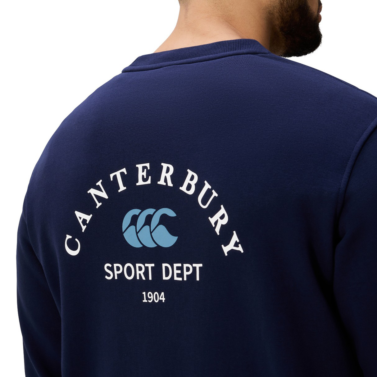 A person wearing the Canterbury Sport Dept. Crew Sweatshirt, a navy blue sweatshirt with "Canterbury Sport Dept 1904" and a blue logo on the back.