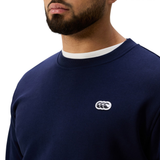 A person wearing the Canterbury Sport Dept. Crew Sweatshirt, athletic fit in navy blue with a white logo on the left chest, layered over a white crew-neck shirt.