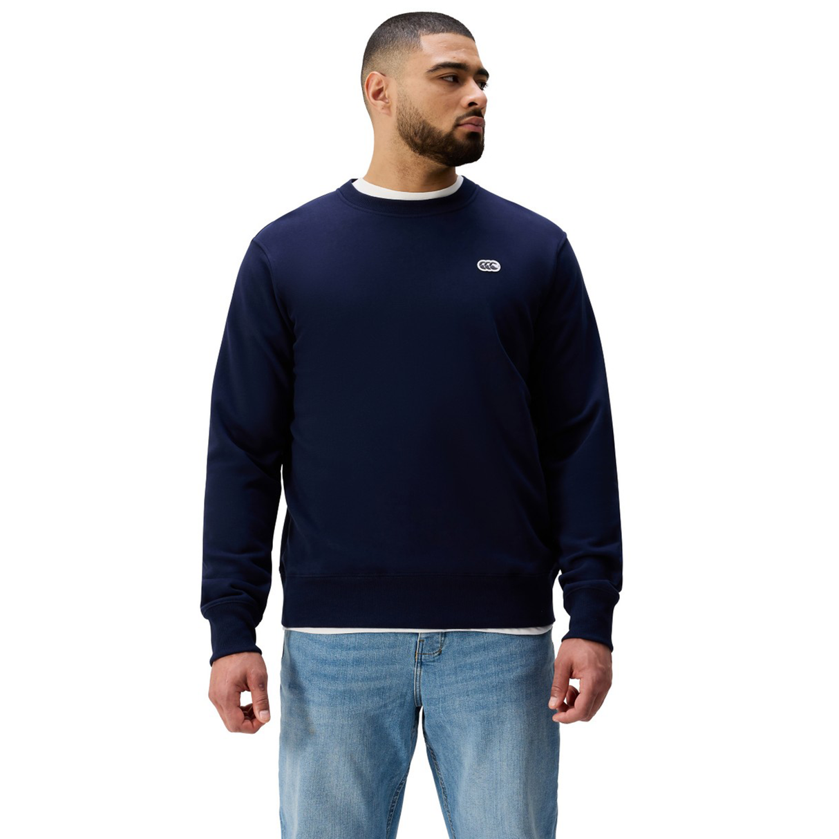 Man in an athletic fit navy Sport Dept. Crew Sweatshirt by Canterbury and light blue jeans, standing with arms relaxed and gazing to the side.