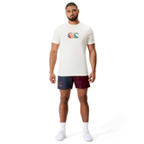 A man faces forward wearing the Canterbury Fundamentals CCC Logo T-Shirt featuring a colorful logo, paired with maroon and navy athletic shorts, white socks, and white sneakers against a plain background.