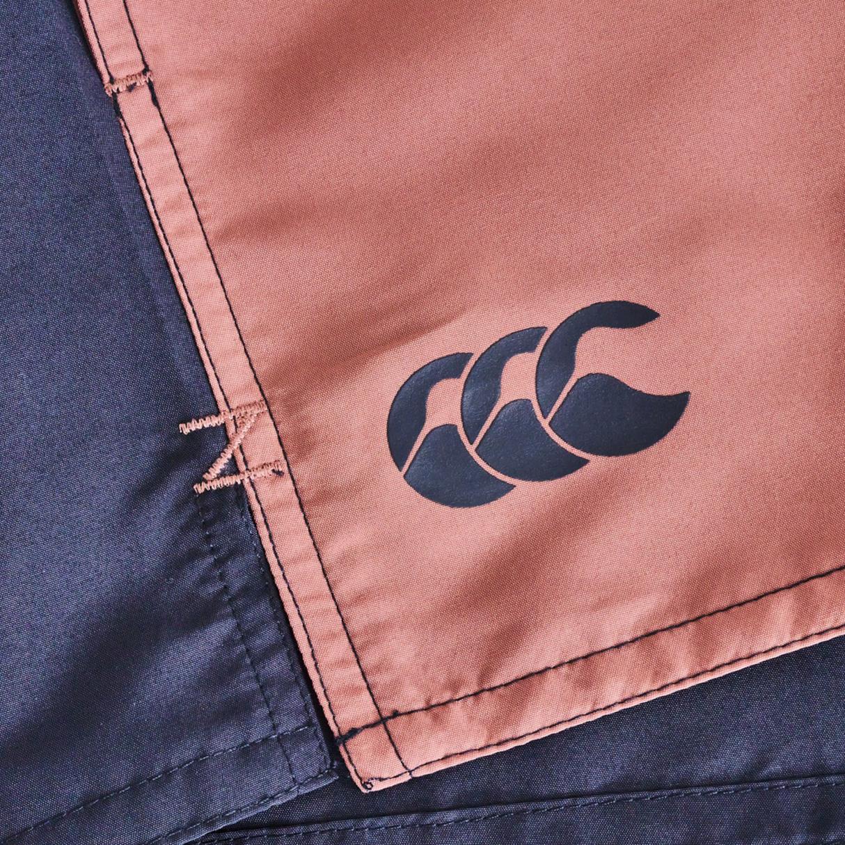 Close-up of Canterbury Women's Fundamentals 3" Harletic Shorts featuring a black Canterbury logo on a brown panel with visible stitching beside a dark blue section—designed for women's rugby and athletic training.