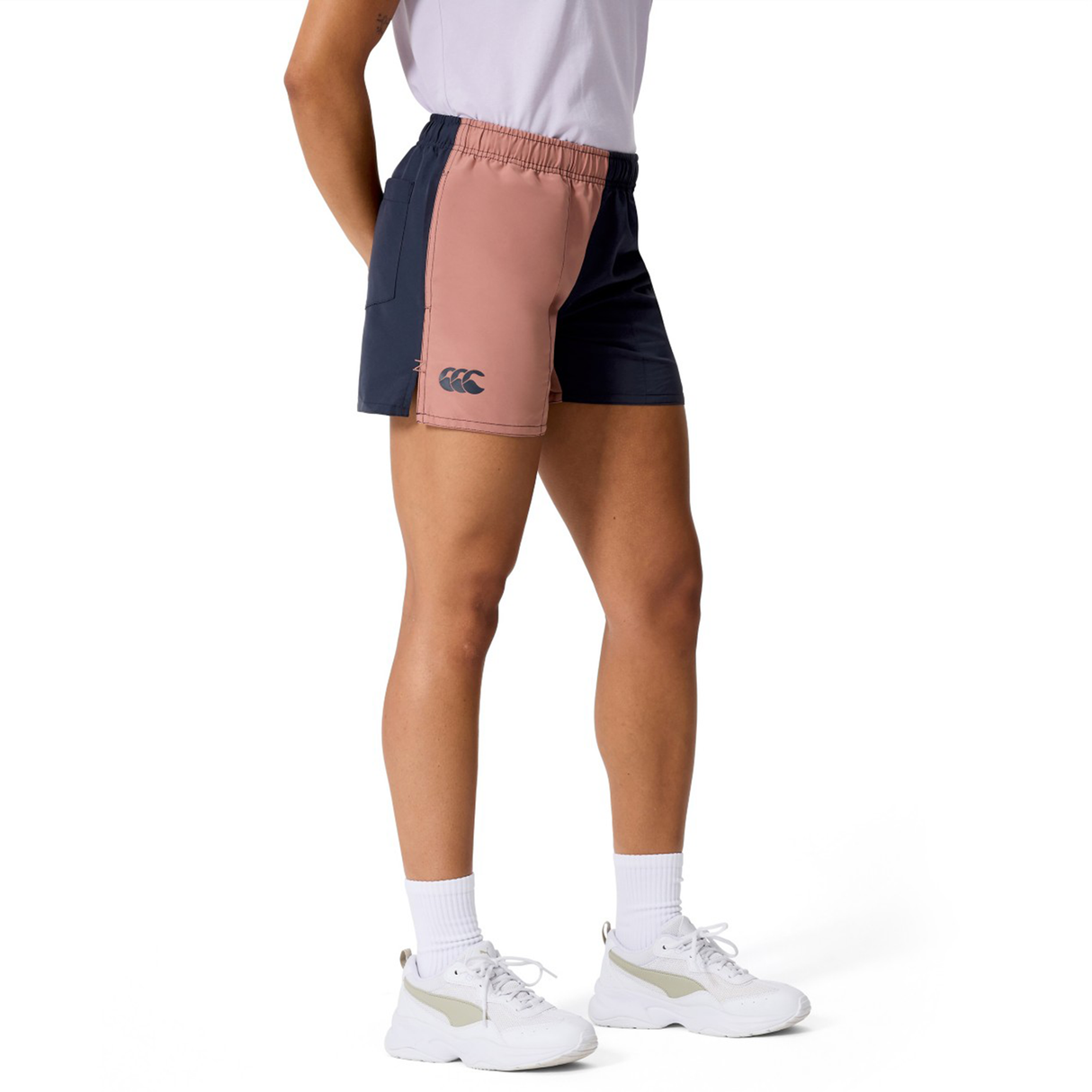 A person wears a white t-shirt, Canterbury Women’s Fundamentals 3" Harletic Shorts in color-blocked pink and navy, white socks, and white sneakers while standing against a plain background.