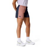 A person wears a white t-shirt, Canterbury Women’s Fundamentals 3" Harletic Shorts in color-blocked pink and navy, white socks, and white sneakers while standing against a plain background.