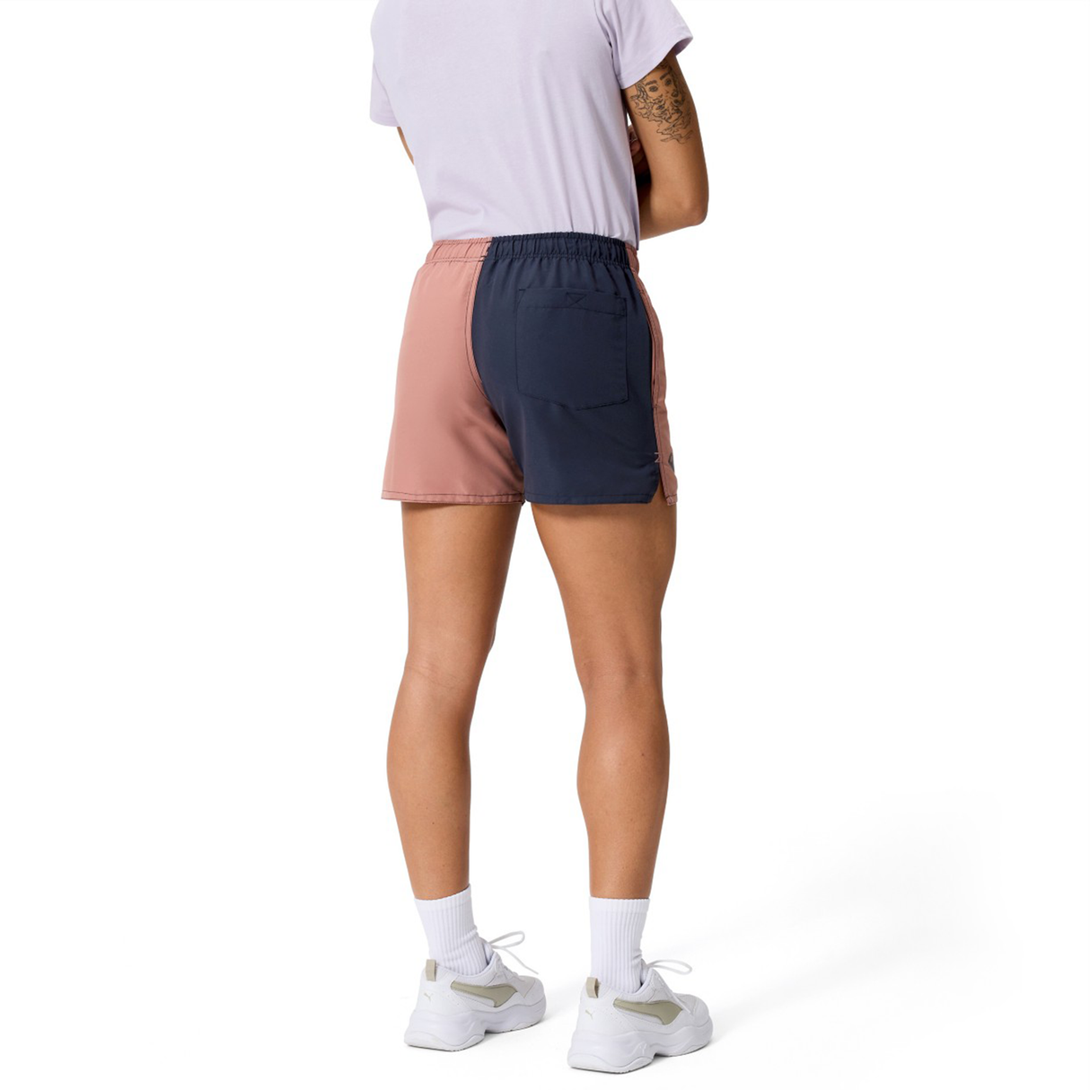 A person stands with their back turned, wearing Canterbury Women's Fundamentals 3" Harletic Shorts in pink and navy, a light purple t-shirt, white socks, and white sneakers.