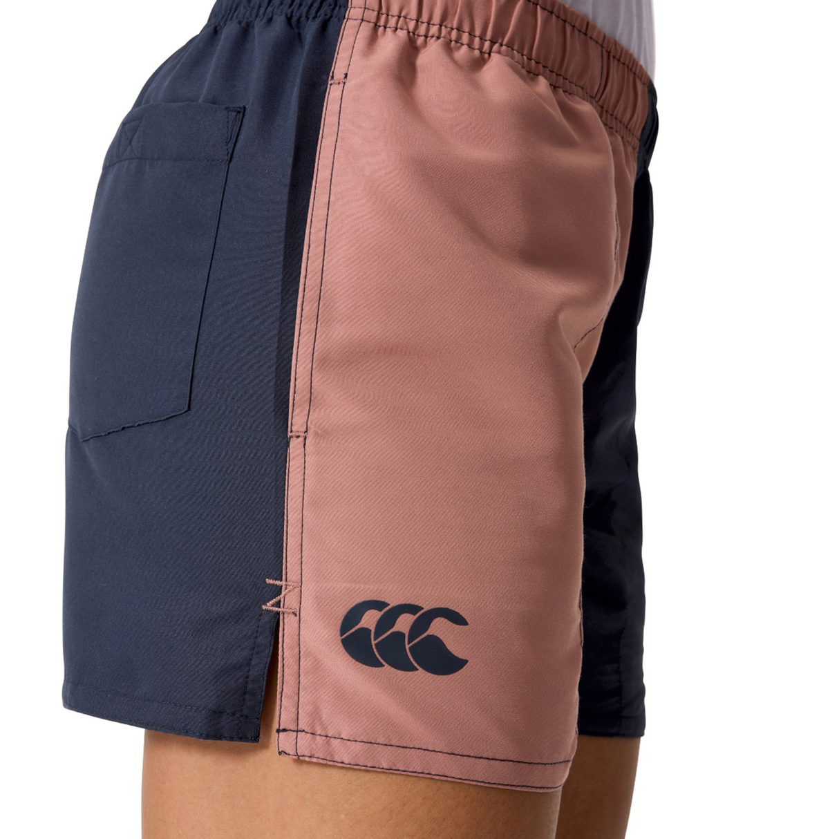 A person wears Canterbury Women's Fundamentals 3" Harletic Shorts featuring navy and pink colorblock panels, a back pocket, and the CCC logo on the left leg.