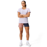 A woman stands with arms crossed, wearing a light purple t-shirt, white socks, white sneakers, and Canterbury Women's Fundamentals 3" Harletic Shorts in pink and navy against a plain white background.