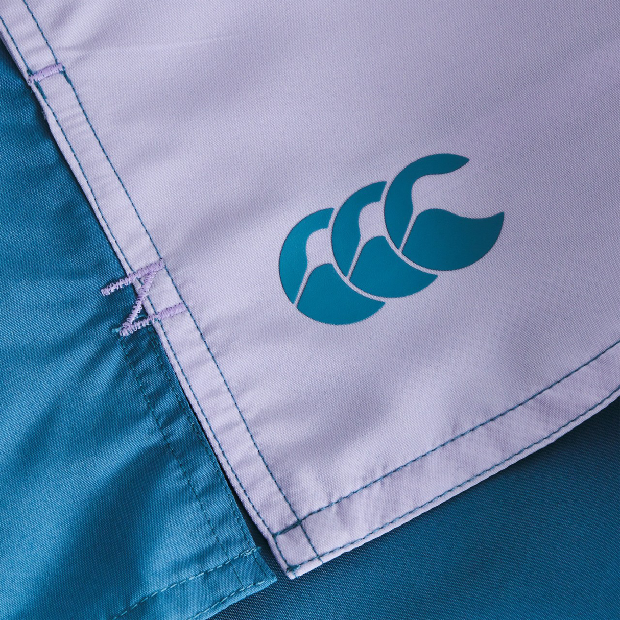 Close-up of the corner of light purple and teal Women's Fundamentals 3" Harletic Shorts by Canterbury, showing stitched seams and a teal logo with three overlapping shapes.