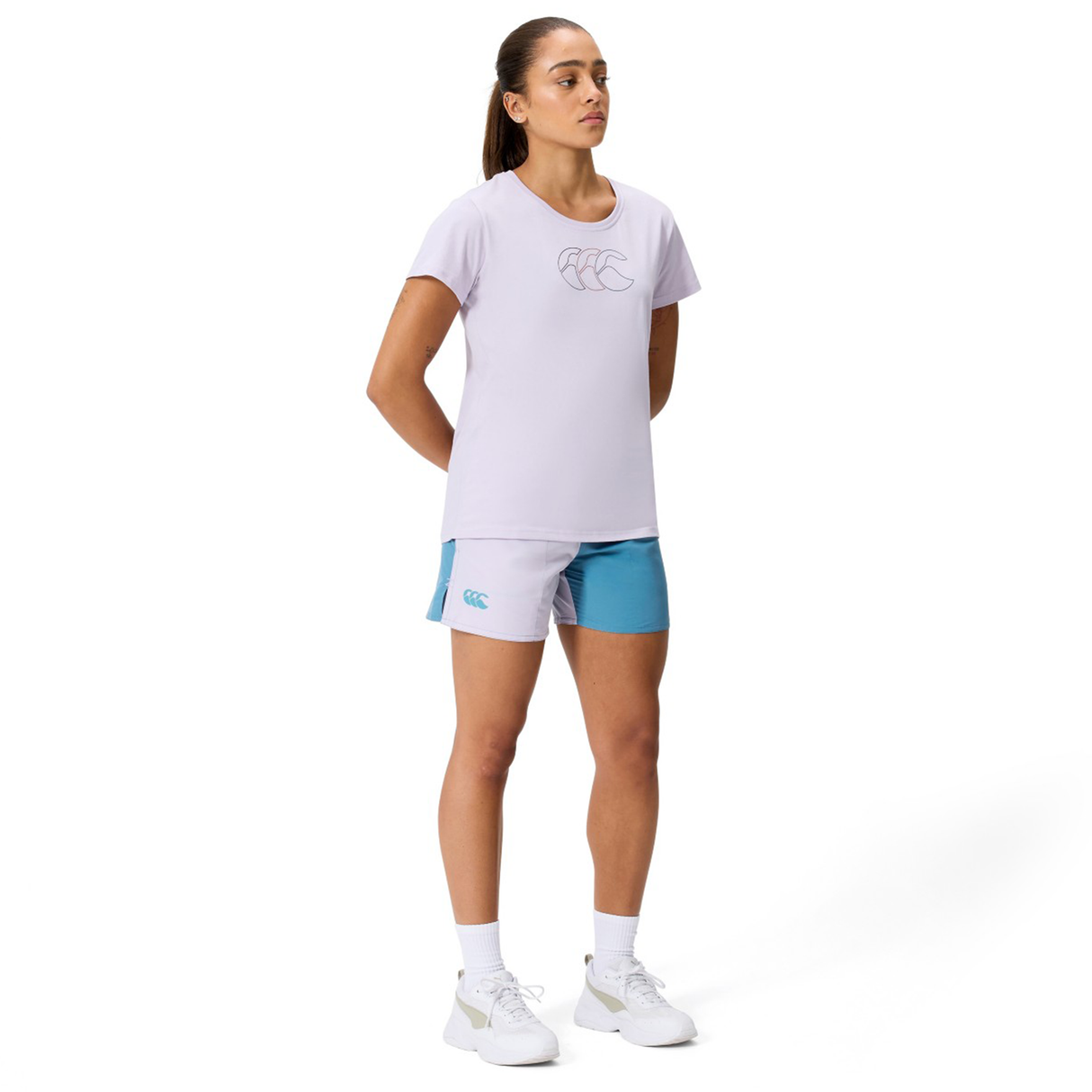 A woman faces forward, wearing a light purple T-shirt, Canterbury Women's Fundamentals 3" Harletic Shorts in light purple and blue, white socks, and white athletic shoes against a plain white background.