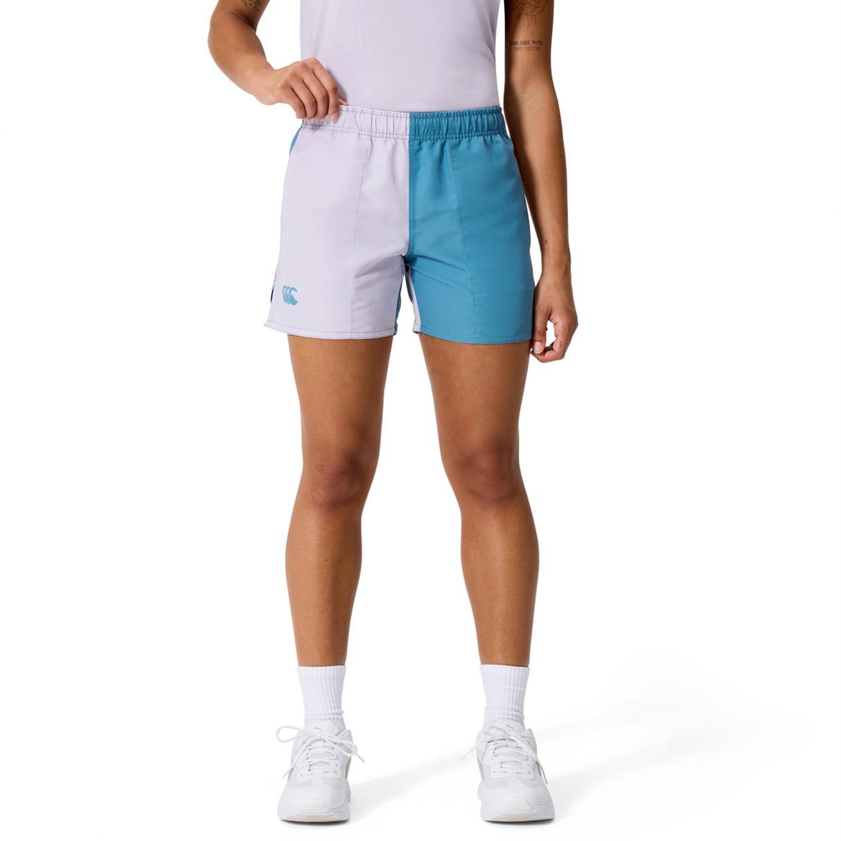 A person stands against a white background wearing Canterbury Women's Fundamentals 3" Harletic Shorts in a white and teal color-block style, paired with a white shirt, socks, and sneakers.