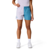 A person stands against a white background wearing Canterbury Women's Fundamentals 3" Harletic Shorts in a white and teal color-block style, paired with a white shirt, socks, and sneakers.