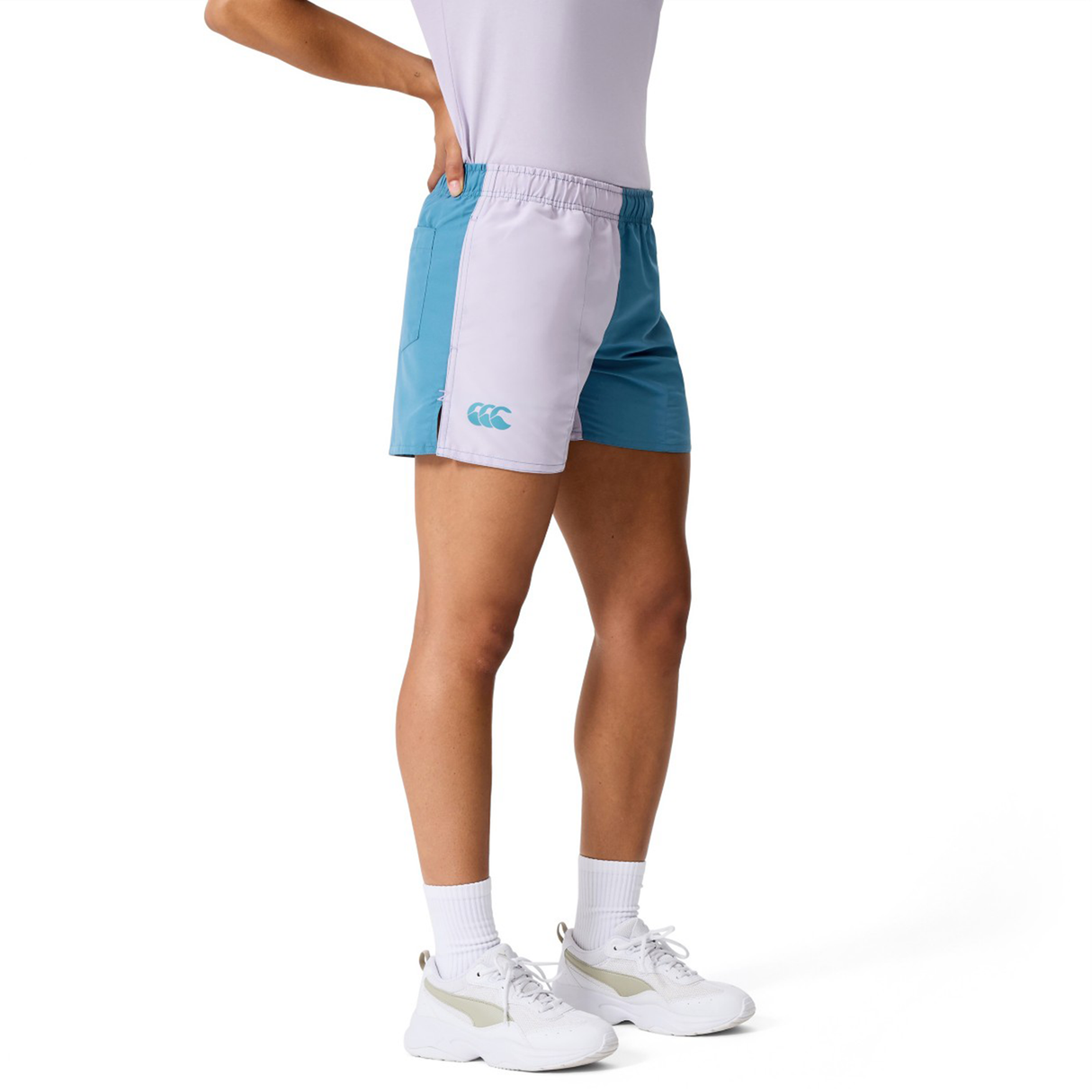 A person wears a white shirt, Women's Fundamentals 3" Harletic Shorts by Canterbury in teal and light gray, white socks, and white sneakers while standing against a plain background.