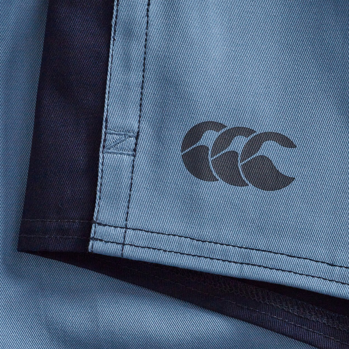 Close-up of the Fundamental Harlequin Drill 3" Shorts by Canterbury in light blue with navy edges, a stitched seam, and black CCC logo—ideal for fans of classic Canterbury rugby shorts.