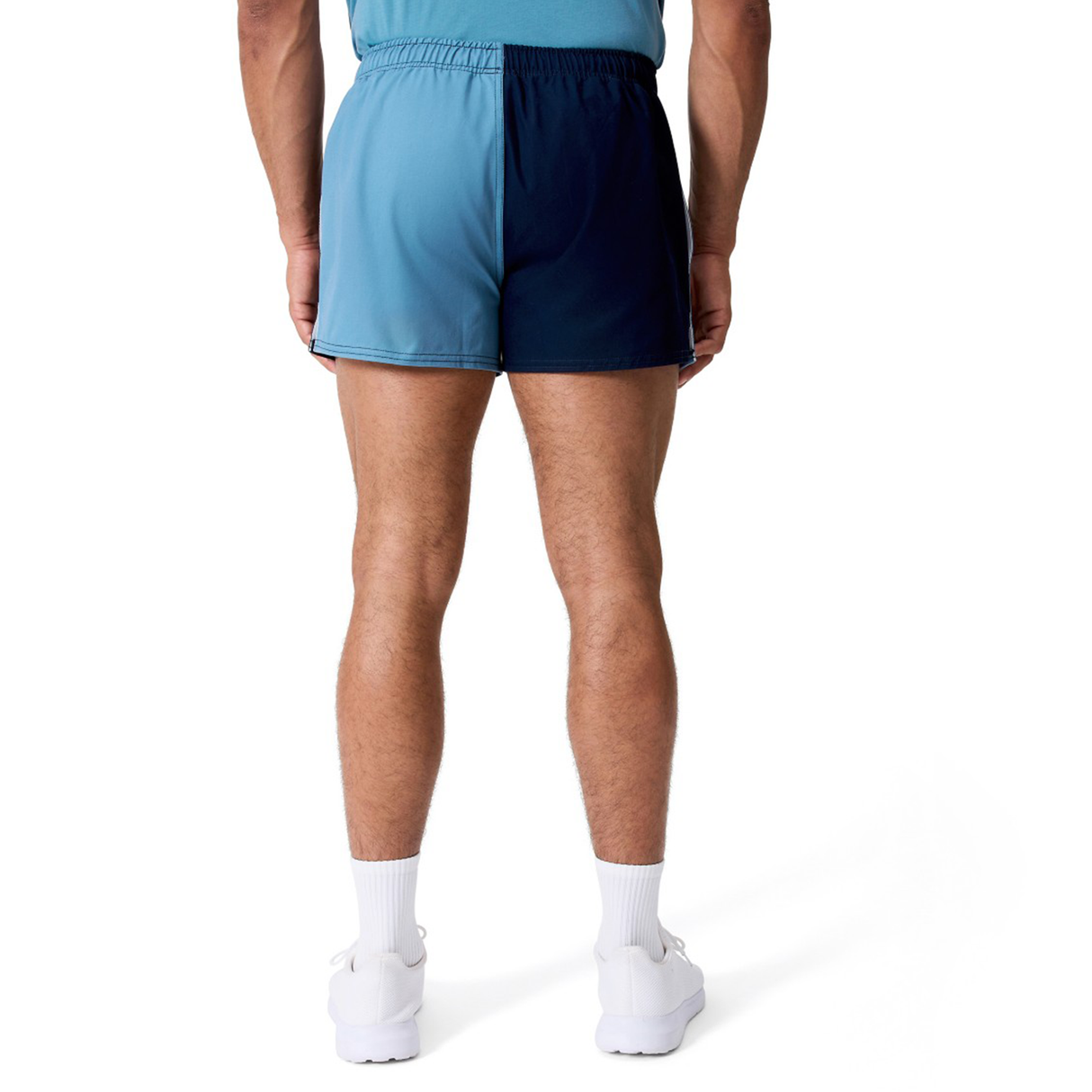 A man stands with his back to the camera wearing Canterbury Fundamental Harlequin Drill 3" Shorts in two-tone blue, paired with white socks and white sneakers, against a plain white background.