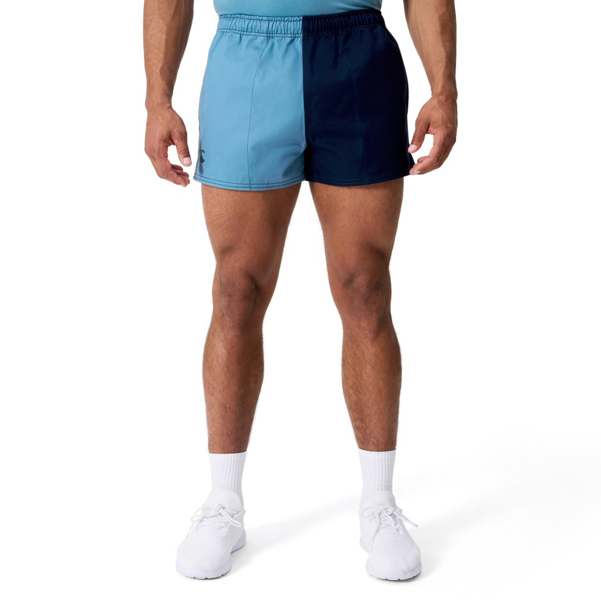 A person wears Canterbury Fundamental Harlequin Drill 3" Shorts in two-tone blue, white socks, and white sneakers, shown from shoulders to feet against a plain background.