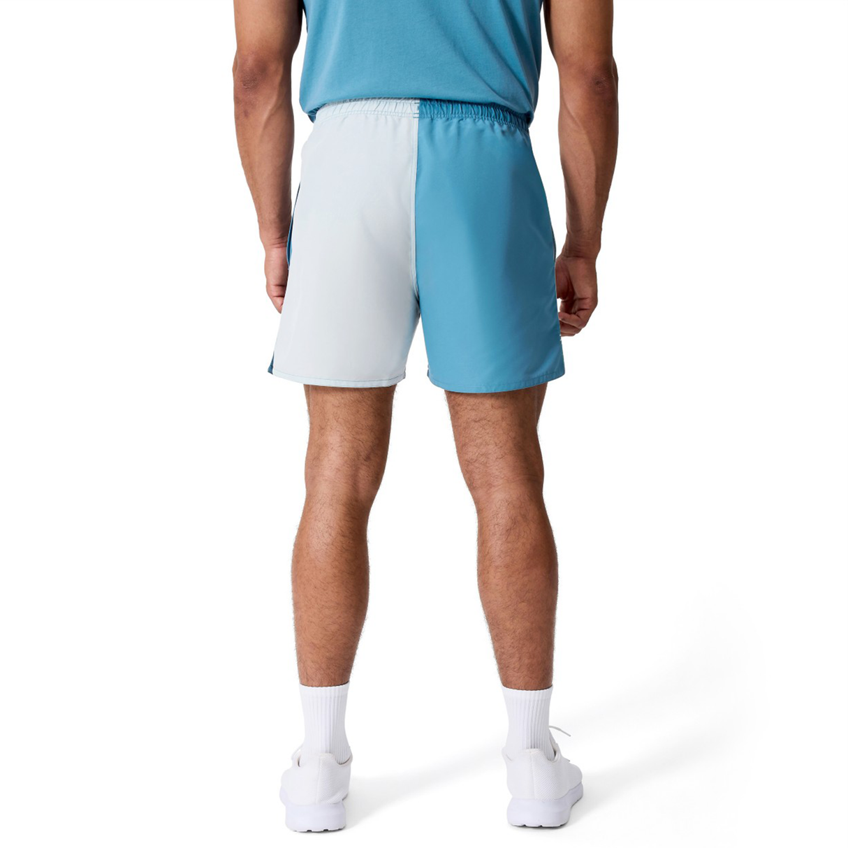 A man stands facing away, wearing a blue shirt, Canterbury Fundamental Harletic 3" Shorts in color-blocked blue and light gray, white socks, and white sneakers against a plain white background.