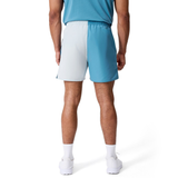 A man stands facing away, wearing a blue shirt, Canterbury Fundamental Harletic 3" Shorts in color-blocked blue and light gray, white socks, and white sneakers against a plain white background.
