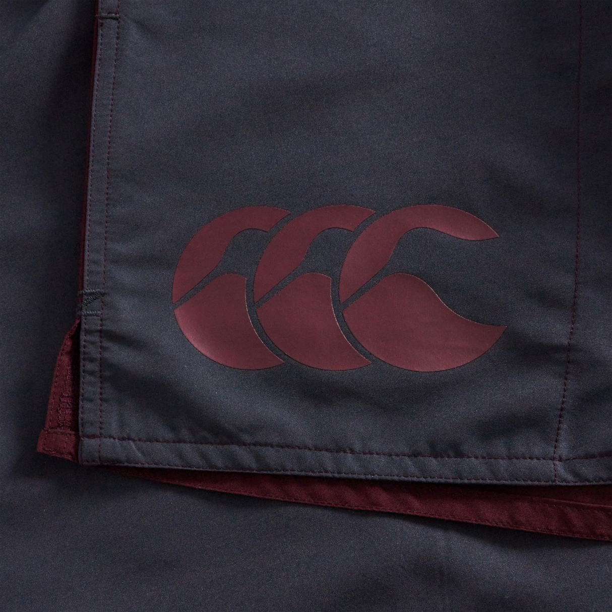 Close-up of Fundamental Harletic 3" Shorts by Canterbury, featuring dark fabric and a maroon Canterbury logo with three overlapping stylized Cs.