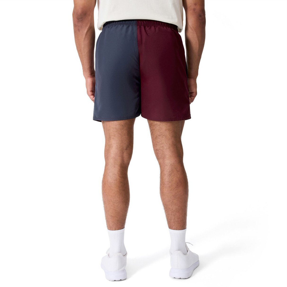 A person stands facing away, wearing Canterbury Fundamental Harletic 3" Shorts with a navy left and burgundy right side, paired with white socks and shoes against a plain white background.