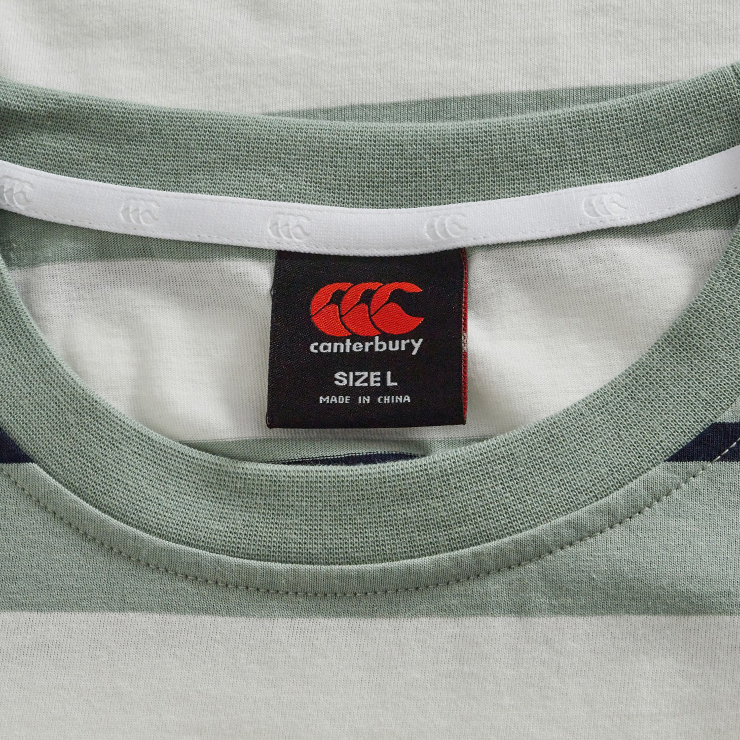 Close-up of a green and white striped collar from the Canterbury Fundamental Yarn Dye Stripe T-Shirt, showing a black brand tag labeled "SIZE L" and "MADE IN CHINA.