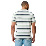 A man faces away, wearing Canterbury's Fundamental Yarn Dye Stripe T-Shirt—a rugby-inspired white short-sleeve tee with black and green stripes—paired with light blue jeans against a plain white background.