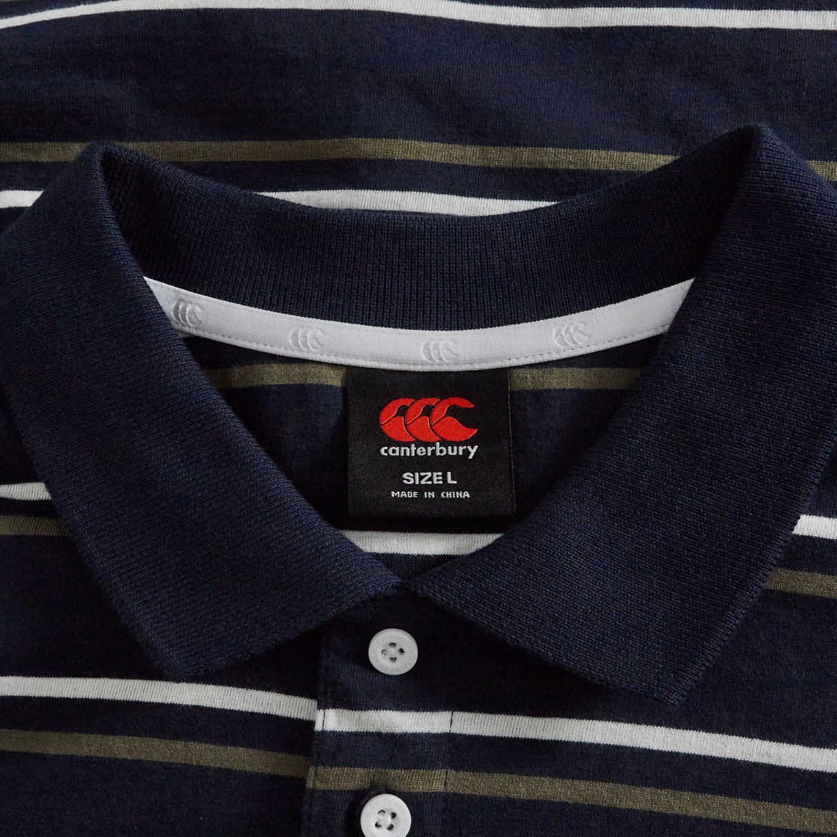 A close-up of the Fundamental Yarn Dye Stripe Polo by Canterbury in navy, showing white buttons and a brand label with size L and "Made in China.