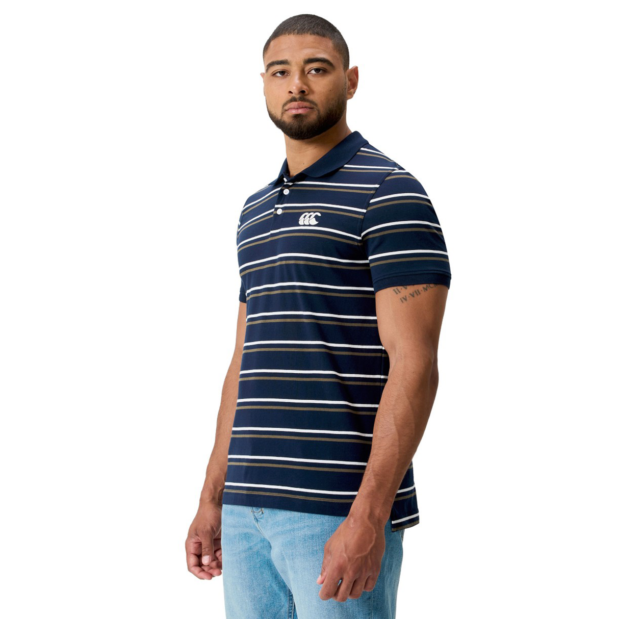 A man in a Canterbury Fundamental Yarn Dye Stripe Polo featuring a white logo on the chest and light blue jeans stands against a plain white background.